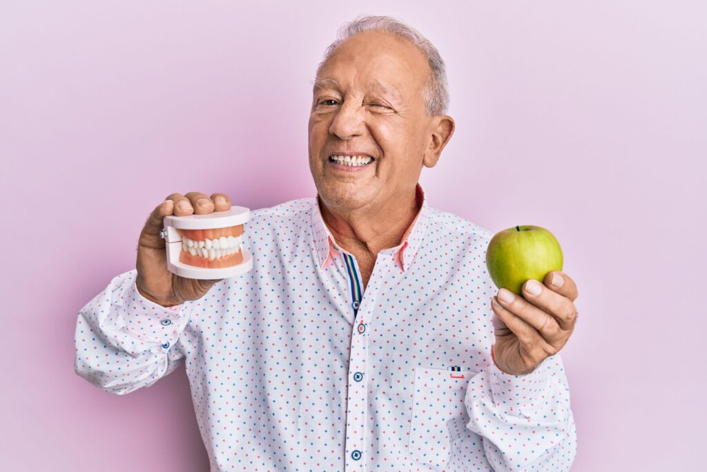 Older man holding green apple and artificial teeth winking with pink background